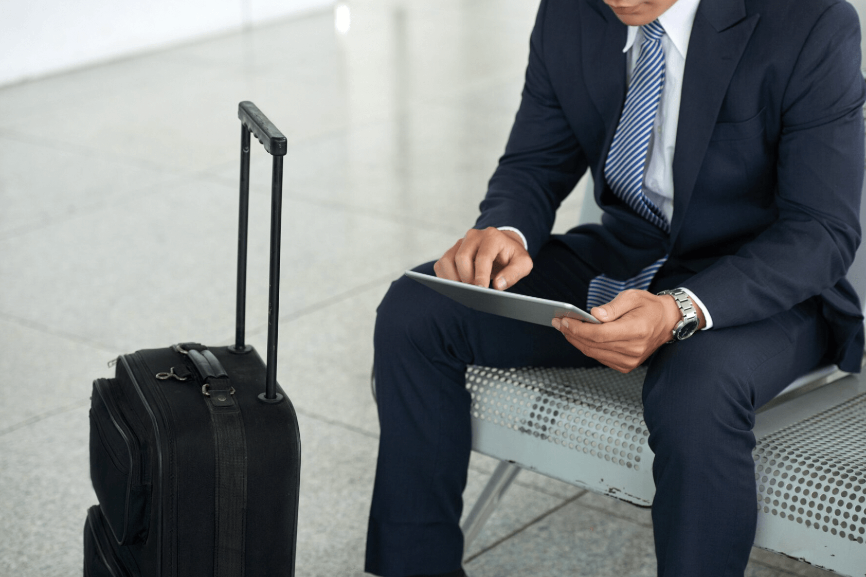 Man in suit seated with tablet and luggage at an airport.